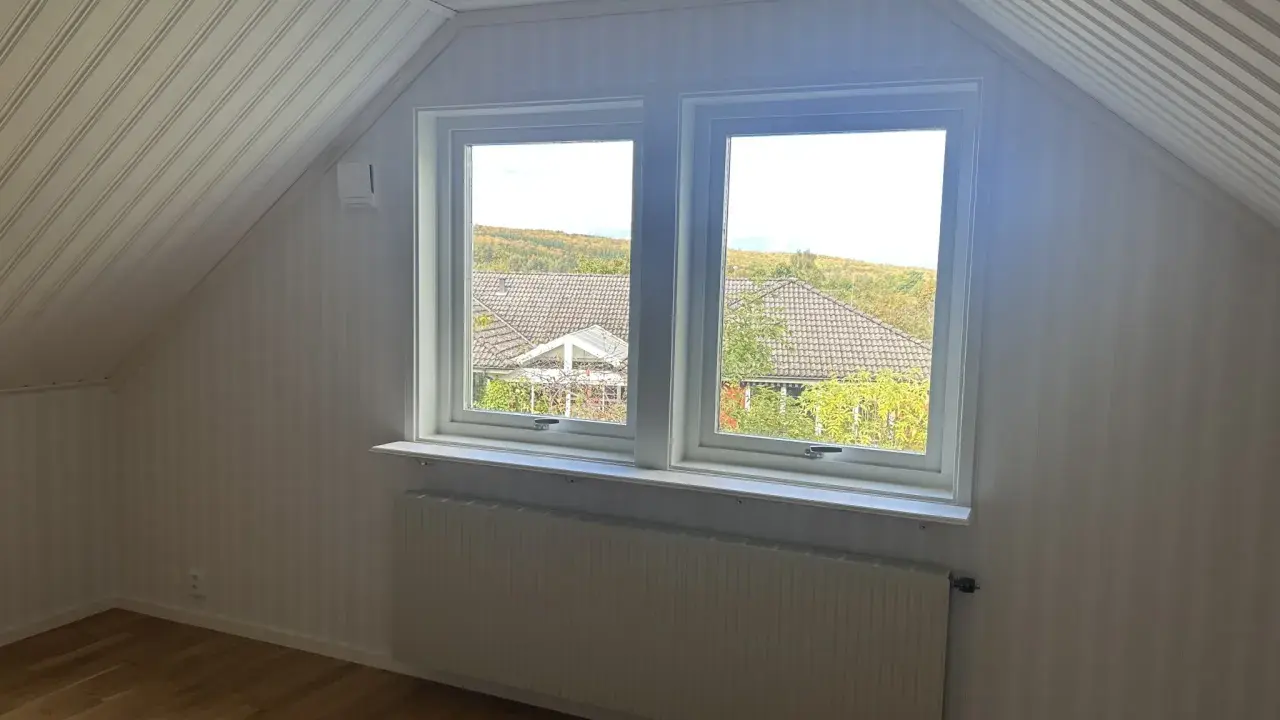 Vitt snedtak med panel och ett fönster med utsikt över träd. Gardiner saknas, men ett element finns under fönstret.