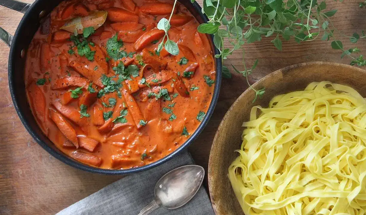En krämig korv stroganoff med morötter i en panna, bredvid en skål med färsk pasta.