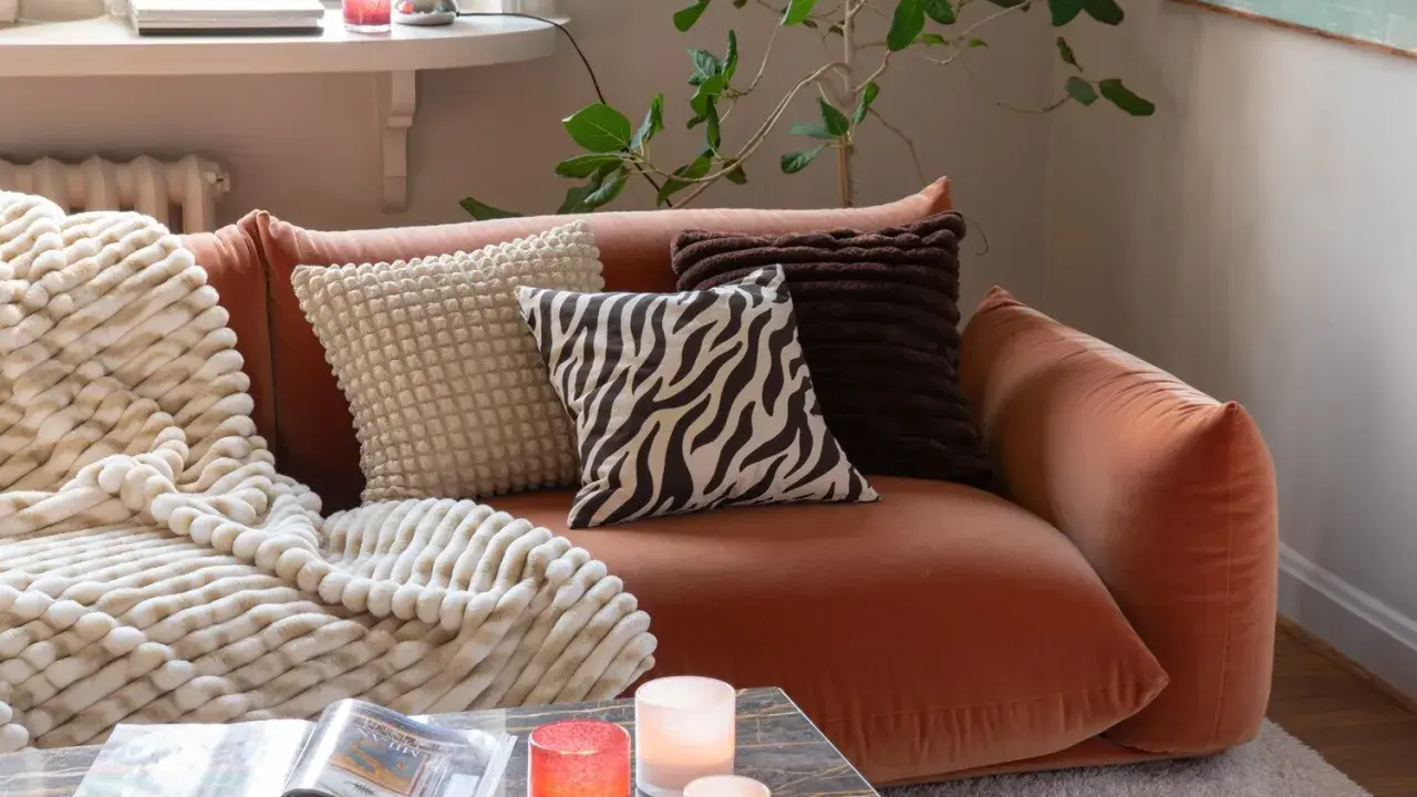 En orange soffa med flera kuddar och en mjuk pläd. Fina dekorationer som ljus och en tidning på soffbordet skapar en mysig atmosfär.