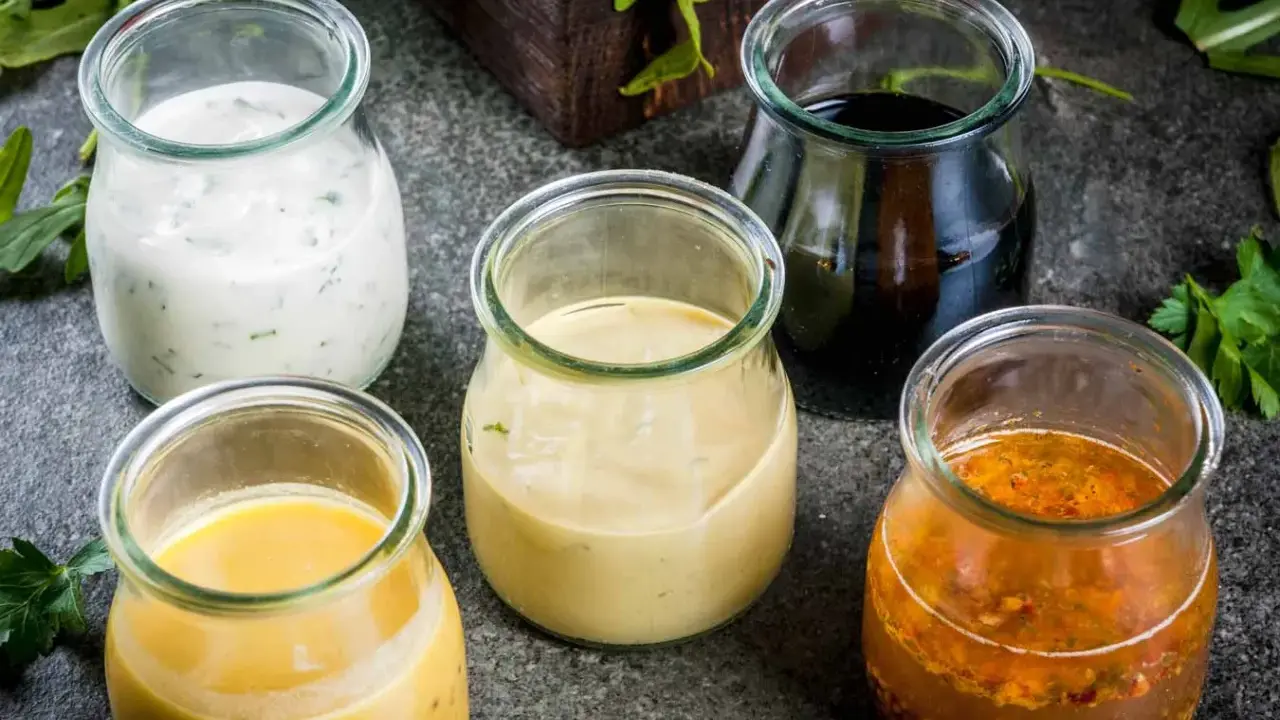 Fem små glasburkar med olika dressing till sallad: krämig vit, gul, ljusbrun, mörk och en med frön.