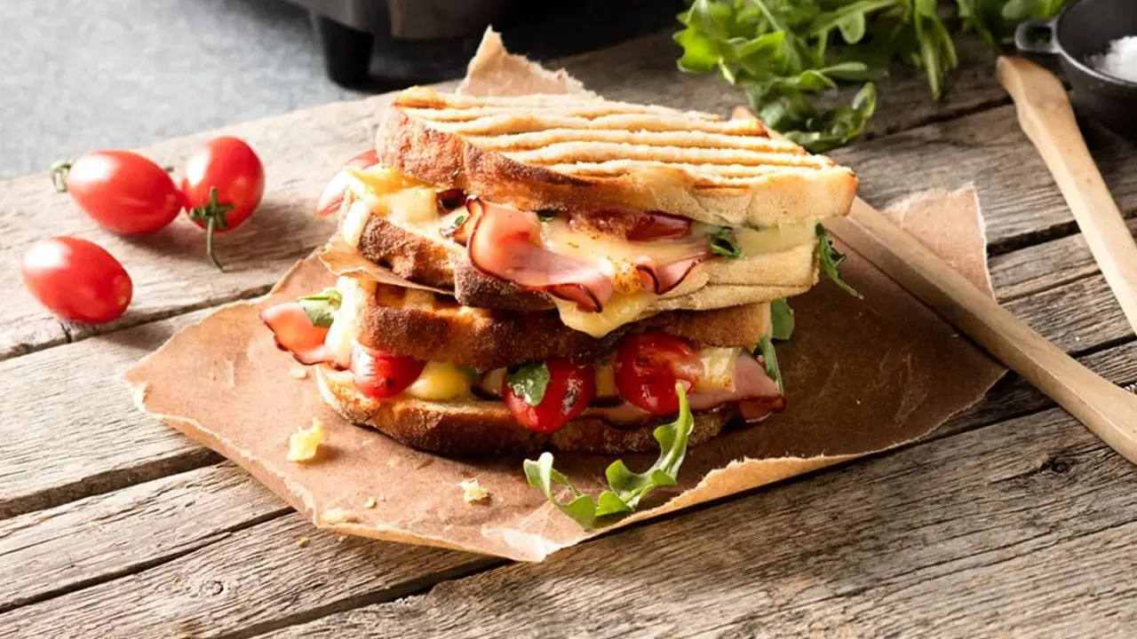 En staplad grilled cheese med skinka, tomater och ruccola på en träyta.