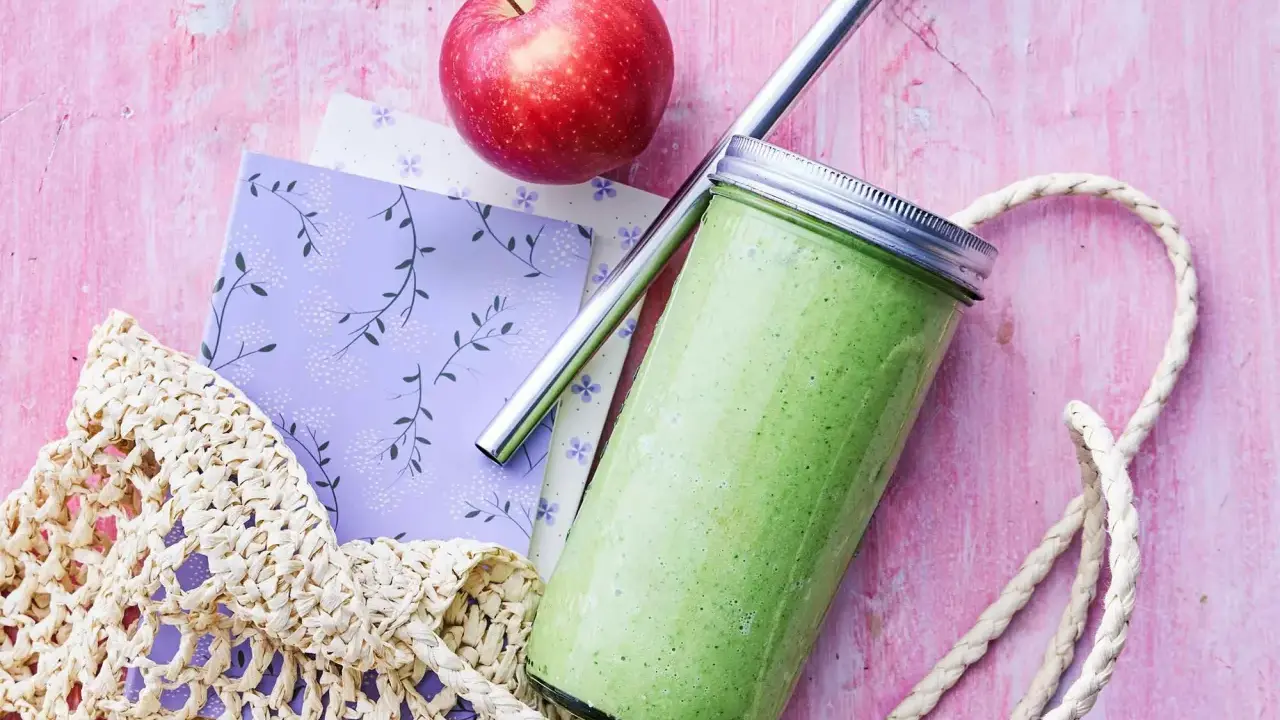 En grön, proteinrik smoothie i glasburk med metallrör, bredvid ett rött äpple och en vävd kasse på en rosa bakgrund.