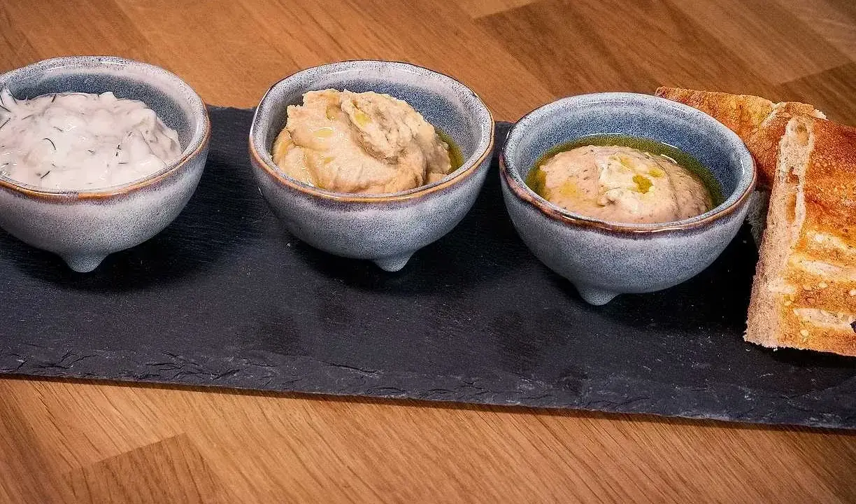 Tre skålar med kall mat, troligen dippsåser eller röror, serveras med rostat bröd på en svart stenplatta.
