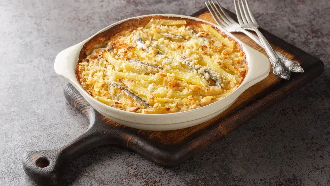 En krämig Janssons frestelse med potatis, ansjovis och ströbröd i en ugnsform, redo att serveras med två silvergafflar.