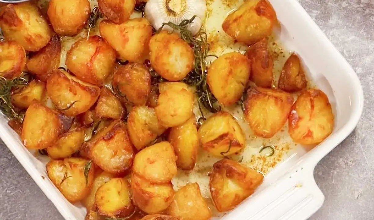 Ugnsrostad potatis i en vit form, gyllene och krispiga, med en hel vitlöksklyfta och rosmarin.