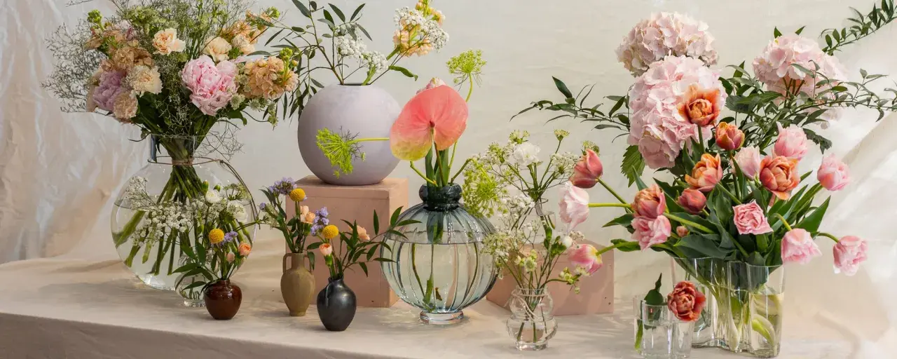 En samling vaser med olika typer av blommor, inklusive pioner, tulpaner och hortensior i mjuka pastellfärger.