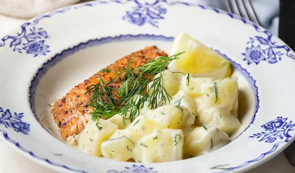 En bit stekt lax med citron och en generös portion krämig dillstuvad potatis, serverad på en vacker blåvit tallrik.
