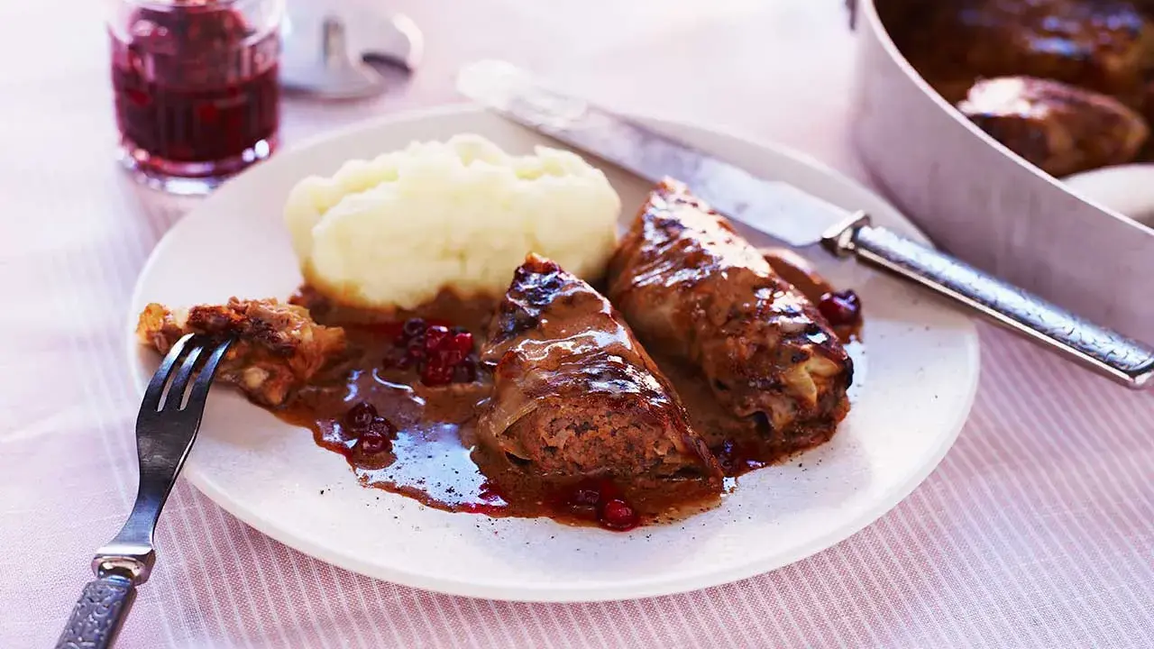 Kåldolmar recept: två kåldolmar med potatismos och lingonsylt, serverade på en tallrik med en gaffel och kniv.