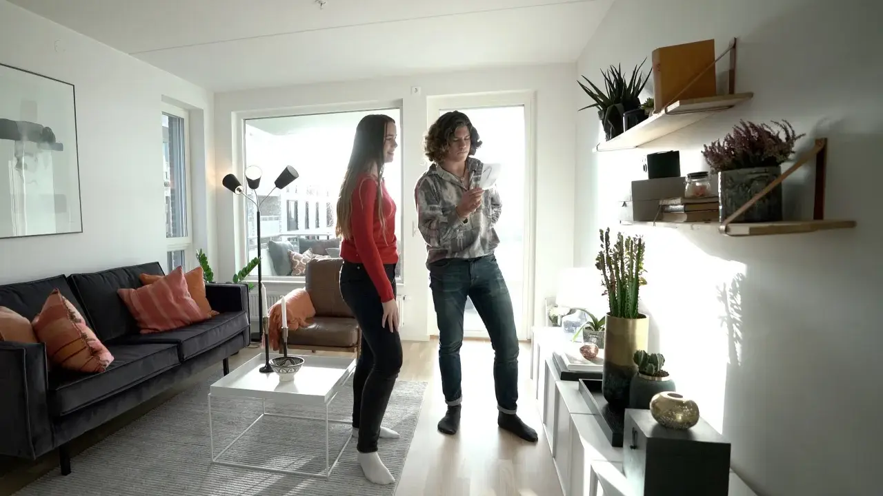Två personer diskuterar något i ett modernt vardagsrum med trendig inredning, mörk soffa och hyllor med växter.