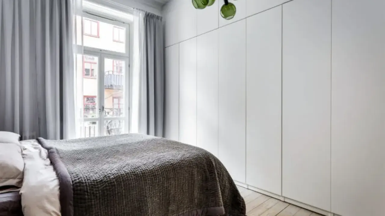 Sovrum med en stor, vit garderob med draperi. En säng med mörkt överkast står framför ett fönster med ljusgrå gardiner.