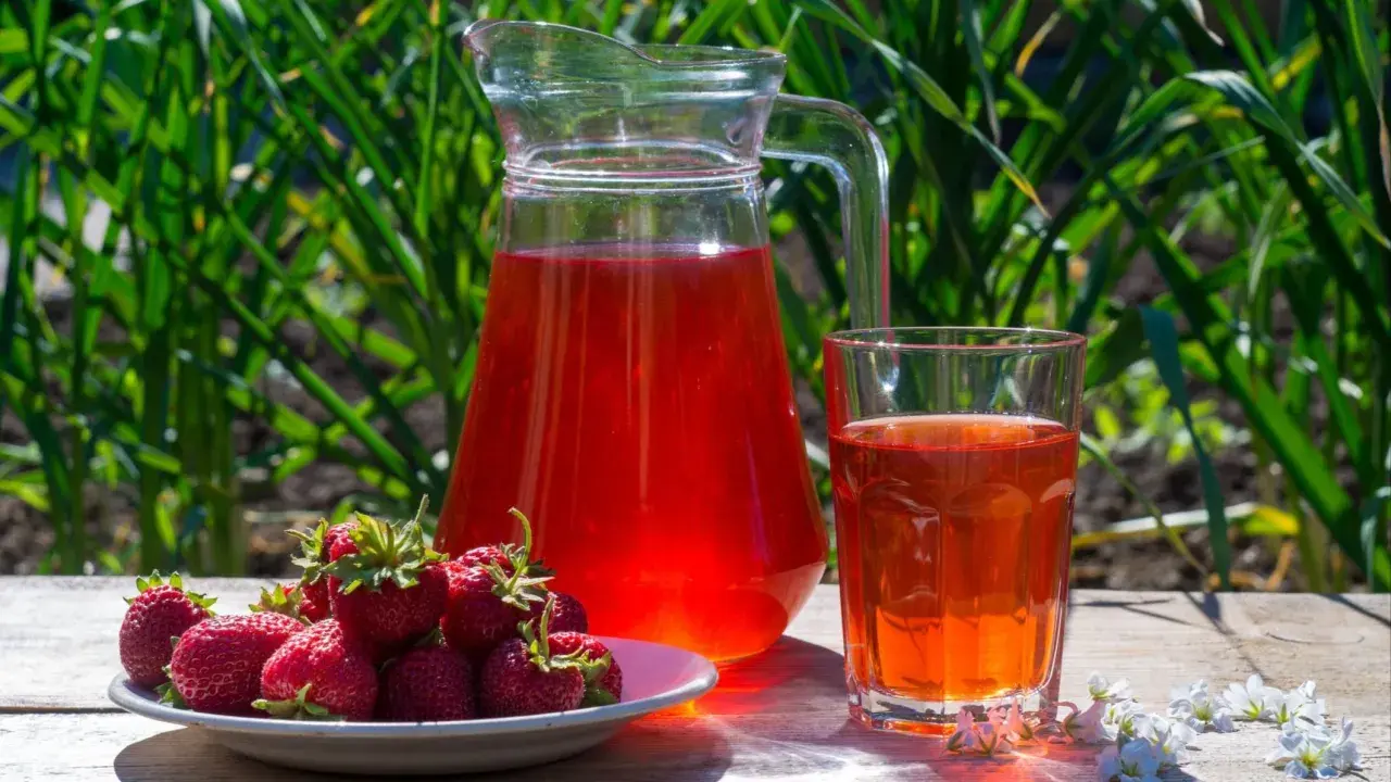 En karaff och ett glas med röd saft står på ett träbord. En tallrik med färska jordgubbar bredvid. Perfekt för ett jordgubbssaft recept.