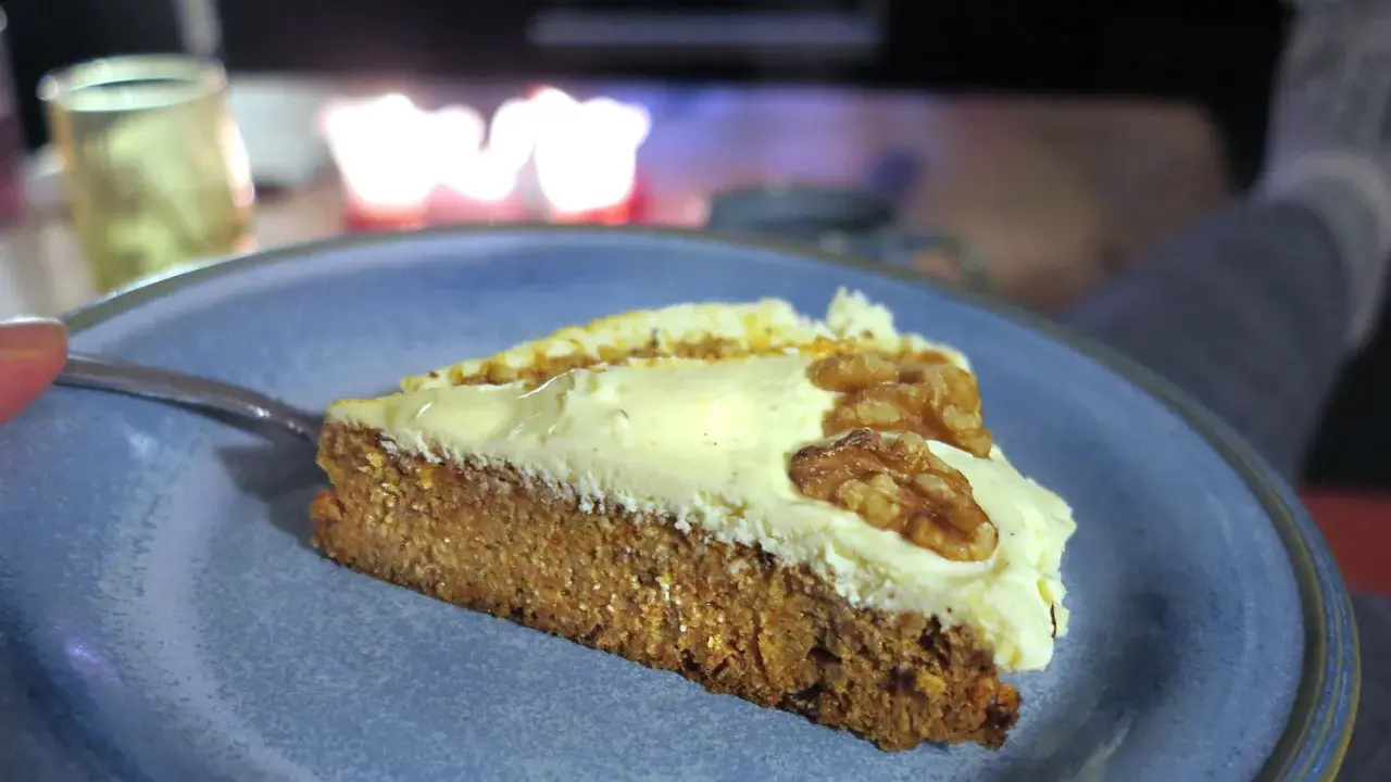 En bit saftig glutenfri morotskaka med krämig frosting och valnötter på en blå tallrik.