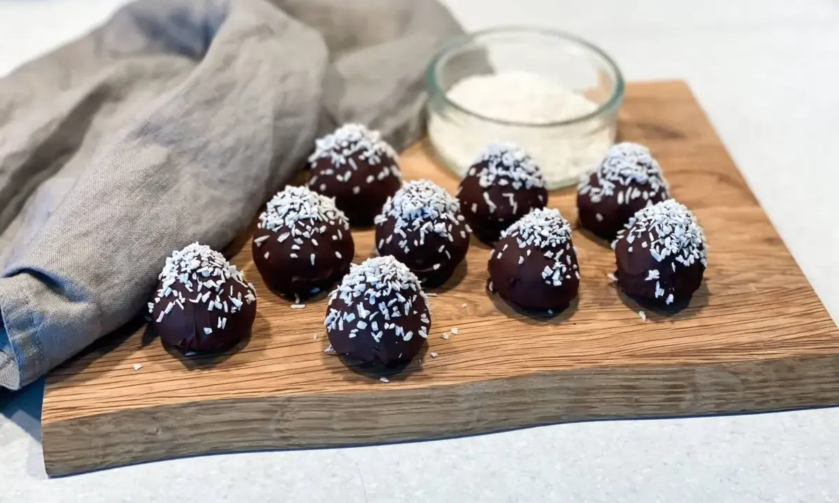 Mörka, chokladöverdragna protein chokladbollar toppade med kokosflingor ligger på en träskärbräda.