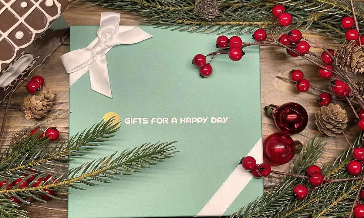 En grön presentask med texten "GIFTS FOR A HAPPY DAY", dekorerad med julgranspynt, kottar och röda bär. Perfekt för julklappar och julrim.