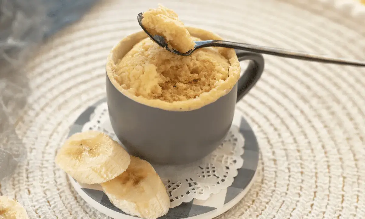 En fluffig mugcake proteinpulver med bananskivor. En sked lyfter en bit av den läckra kakan.