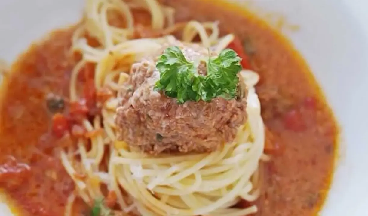En portion spaghetti med en stor frikadell i en krämig tomatsås, toppad med färsk persilja.