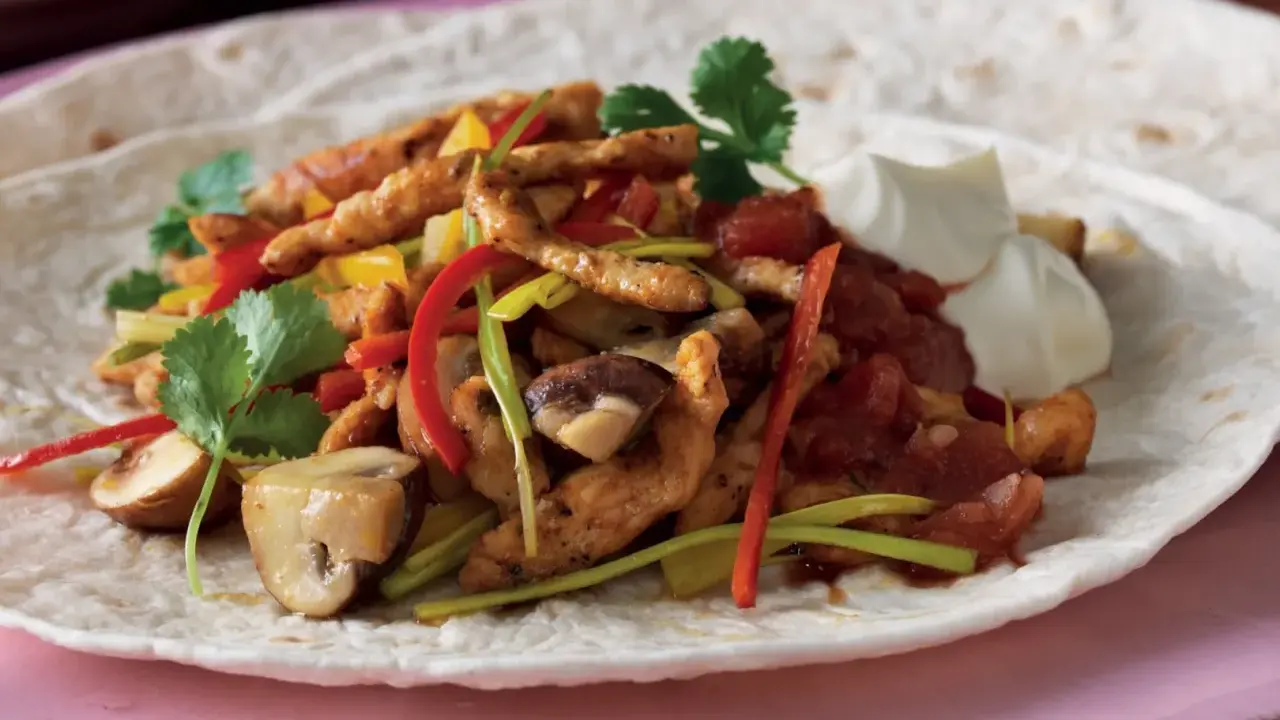 En portion fajitas med kyckling, paprika, champinjoner och lök på en tortilla, serverad med gräddfil. Ett gott fajitas recept!