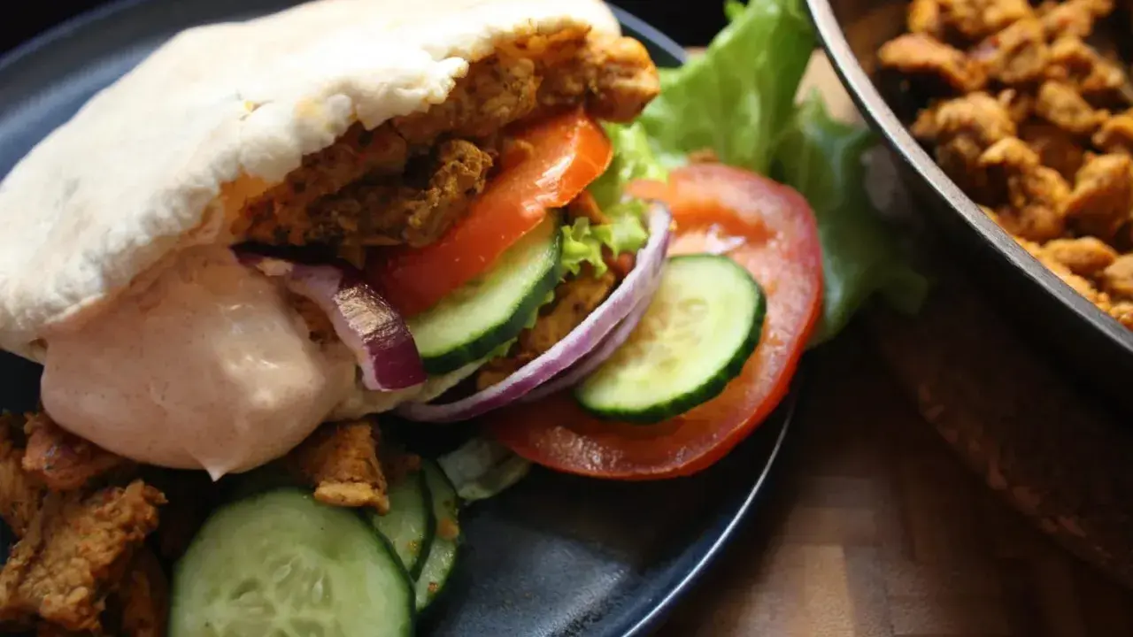 En saftig kyckling kebab i pitabröd med grönsaker och sås. Bredvid ligger mer kyckling i en panna.