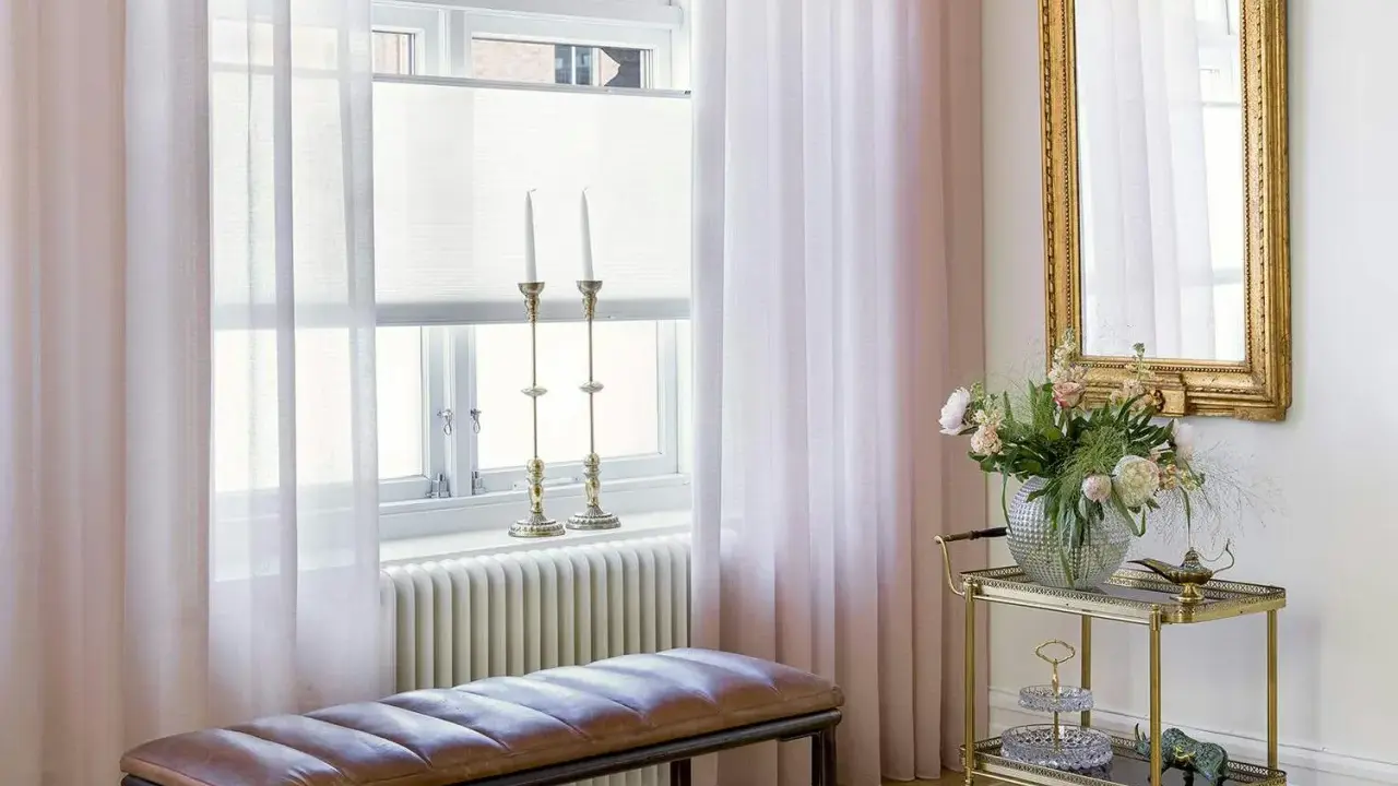 Ljusrosa gardiner längd från golvet ramar in ett fönster med ljusinsläpp. En brun läderbänk står framför ett element.