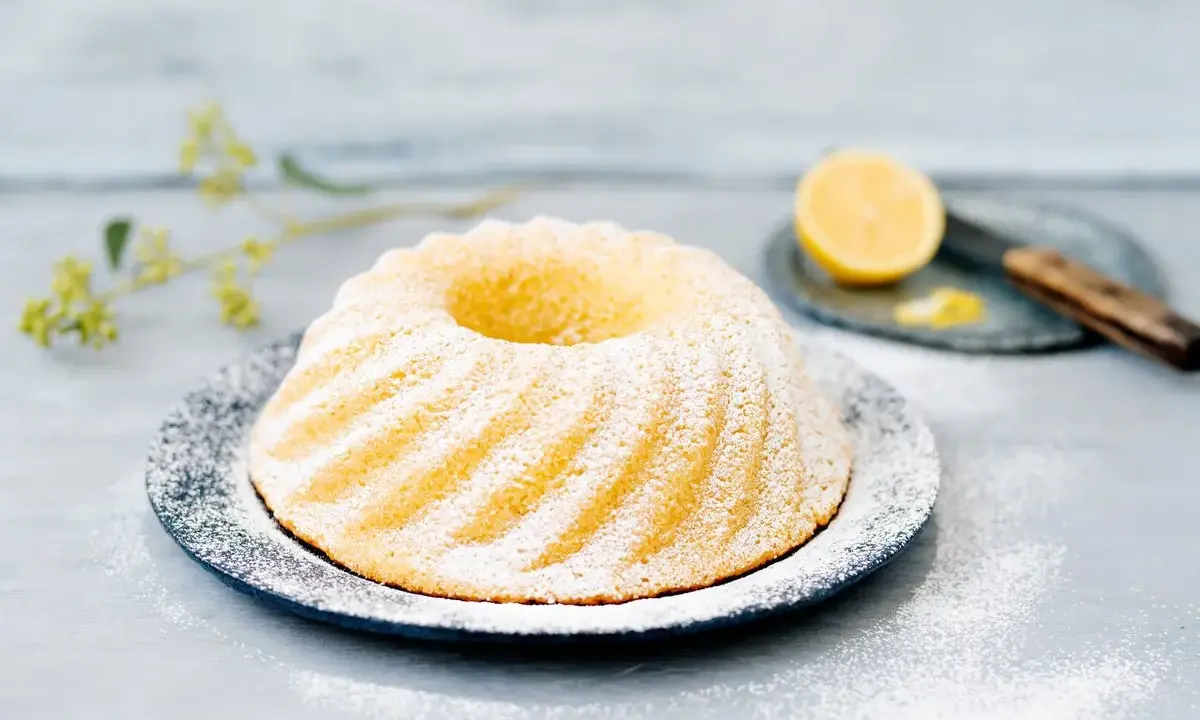 En vacker citronkaka i långpanna, pudrad med florsocker, står på ett fat. En halv citron och en kniv syns i bakgrunden.