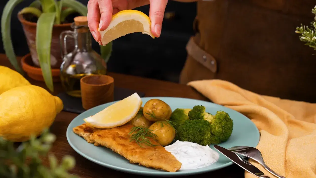 En hand pressar citron över en tallrik med panerad torsk, potatis, broccoli och en klick sås.