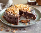 En saftig kladdkaka med kokostosca, toppad med rostad kokos och chokladglasyr. En sked med chokladsås ligger bredvid.