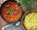 En krämig korv stroganoff med morötter i en panna, bredvid en skål med färsk pasta.