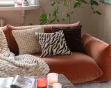 En orange soffa med flera kuddar och en mjuk pläd. Fina dekorationer som ljus och en tidning på soffbordet skapar en mysig atmosfär.