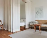 Vita draperier skärmar av ett rum och skapar en mysig sovalkov. En trästol och en soffa med kuddar står i rummet.