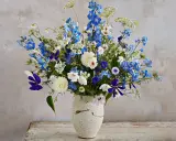 En vacker bukett med blå och vita perenna snittblommor, som riddarsporre och prästkrage, i en rustik vas.