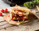 En staplad grilled cheese med skinka, tomater och ruccola på en träyta.
