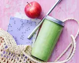 En grön, proteinrik smoothie i glasburk med metallrör, bredvid ett rött äpple och en vävd kasse på en rosa bakgrund.