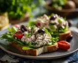 Två smörgåsar med tonfisk sallad, ruccola, tomater och oliver på en tallrik.
