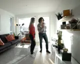 Två personer diskuterar något i ett modernt vardagsrum med trendig inredning, mörk soffa och hyllor med växter.
