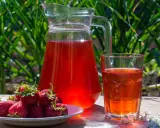 En karaff och ett glas med röd saft står på ett träbord. En tallrik med färska jordgubbar bredvid. Perfekt för ett jordgubbssaft recept.