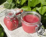 Två glasburkar med hemgjord rabarbermarmelad står på en träbänk utomhus, omgivna av gröna blad.