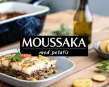 En väldoftande moussaka med potatis, serverad i en ugnsform. Rätten är dekorerad med basilika och tomater.