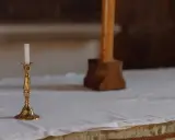 En ljusstake med ett vitt ljus står på ett vitt dukat bord. En träkorsfot skymtar i bakgrunden. Perfekt dop inspiration.