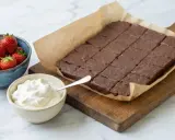 En kladdkaka i långpanna, uppskuren i rutor, serveras med vispad grädde och färska jordgubbar. Perfekt för fikat!