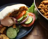 En saftig kyckling kebab i pitabröd med grönsaker och sås. Bredvid ligger mer kyckling i en panna.