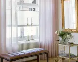 Ljusrosa gardiner längd från golvet ramar in ett fönster med ljusinsläpp. En brun läderbänk står framför ett element.