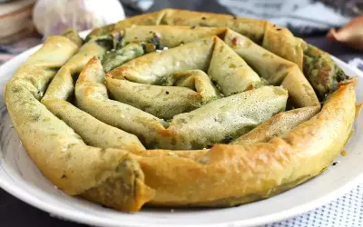 En gyllene, spiralformad burek med spenatfyllning, perfekt för ett hemlagat burek recept. Vitlök och lök i bakgrunden.