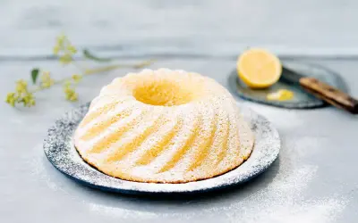 En vacker citronkaka i långpanna, pudrad med florsocker, står på ett fat. En halv citron och en kniv syns i bakgrunden.