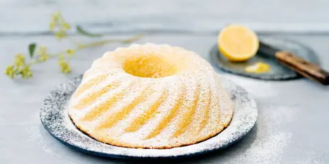 En saftig sockerkaka citron, pudrad med florsocker, serveras på ett fat. En halv citron och en kniv syns i bakgrunden.