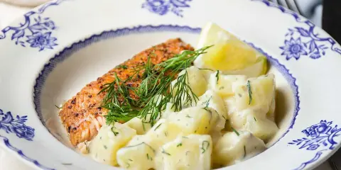 En bit stekt lax med citron och en generös portion krämig dillstuvad potatis, serverad på en vacker blåvit tallrik.