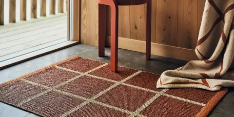 En rödbrun Pappelina Leksand-matta med rutmönster, en orange pall och en filt med randigt mönster i en mysig miljö.