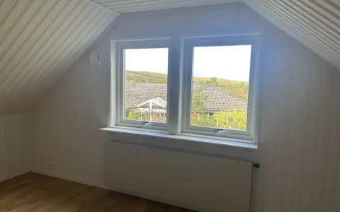Vitt snedtak med panel och ett fönster med utsikt över träd. Gardiner saknas, men ett element finns under fönstret.