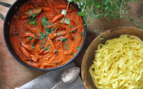 En krämig korv stroganoff med morötter i en panna, bredvid en skål med färsk pasta.