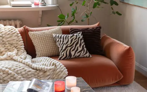 En orange soffa med flera kuddar och en mjuk pläd. Fina dekorationer som ljus och en tidning på soffbordet skapar en mysig atmosfär.