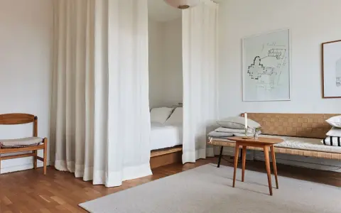 Vita draperier skärmar av ett rum och skapar en mysig sovalkov. En trästol och en soffa med kuddar står i rummet.